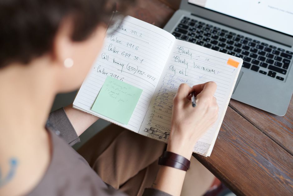 A person writing notes in a notebook beside an open laptop, planning an edit